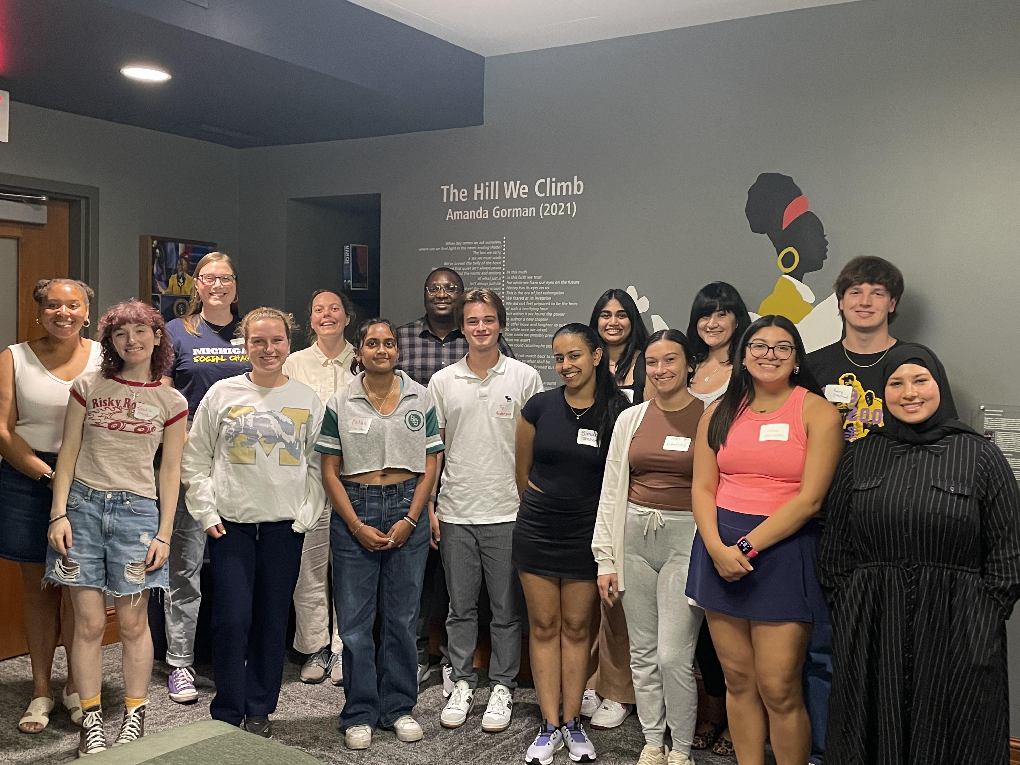 A photo of the Ginsberg Student Advisory Board standing in the Abeng Multicultural Lounge in front of a grey wall with an Amanda Gorman poem behind them. 