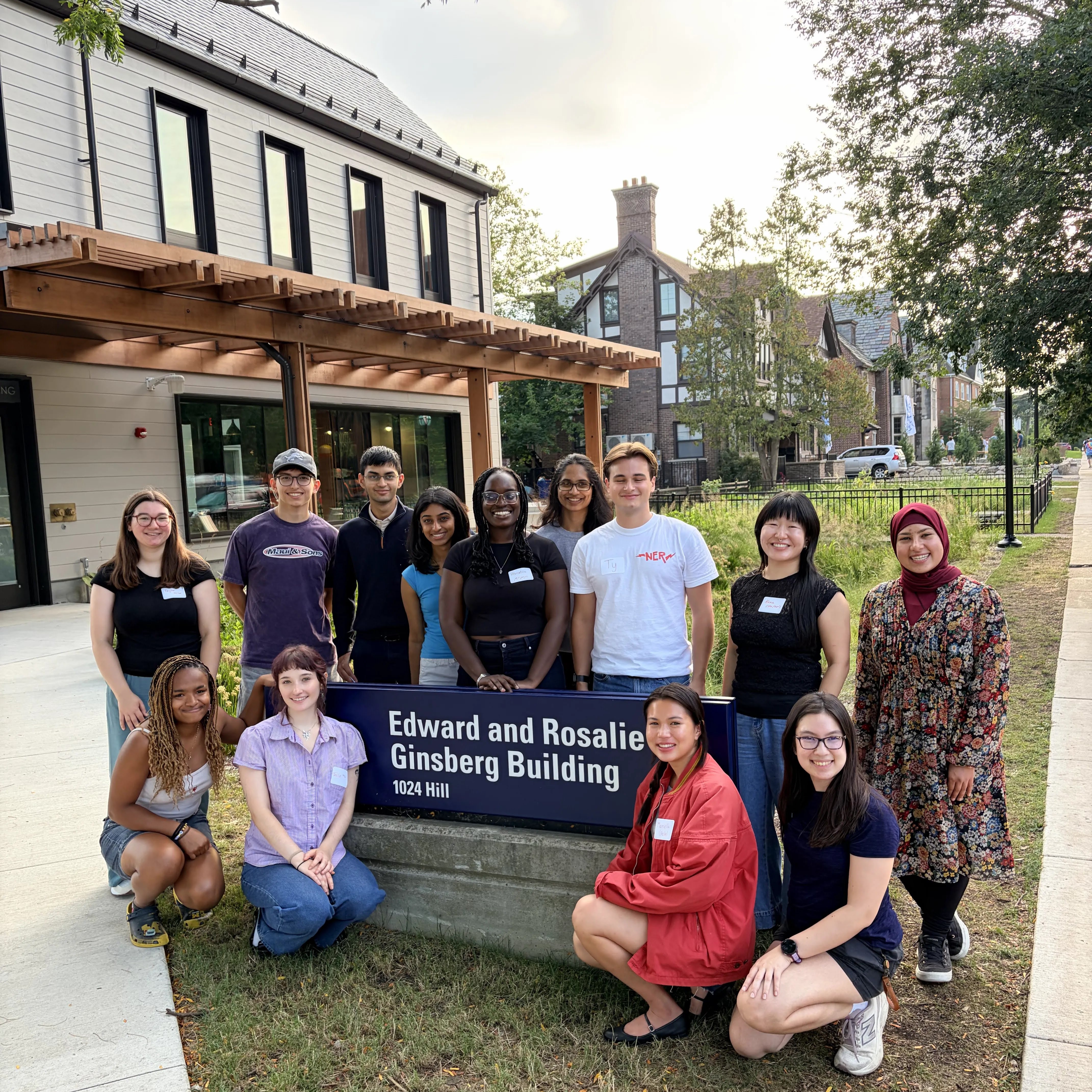 12 students gathered in front of a 2 story building under a blue sky, around a blue sign with the words edward & rosalie ginsberg building