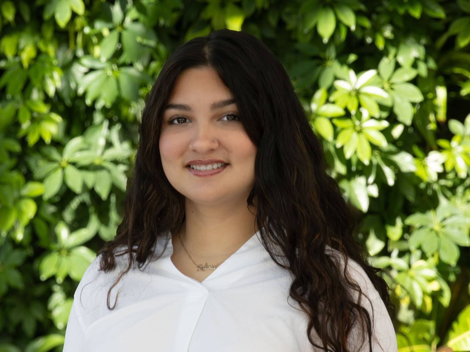 photo of a woman with long dark hair smiling at the camera with green plants behind her