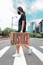 dark haired woman with mask with Justice sign walking along a crosswalk