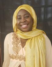 A head-and-shoulders picture of a woman in a golden headscarf. She is wearing a patterned light yellow top and is smiling.