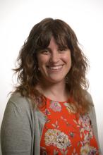 A head-and-shoulders picture of a woman with shoulder-length brown hair. She is wearing a grey cardigan and an orange patterened shirt, and is smiling.