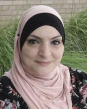 A head-and-shoulders picture of a woman with a pink and black headscarf. She is wearing a patterned top and is smiling gently.