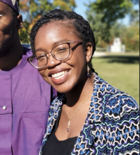 A head-and-shoulders picture of a woman with dark hair pulled back into braids. She is wearing a patterned sweater and glasses, and smiling.