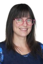 A head-and-shoulders picture of a woman with shoulder-length dark brown hair and glasses. She is wearing a dark blue top and is smiling.