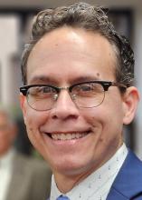 A close-up picture of a man with short brown hair and glasses. He is wearing a collared shirt and is smiling.