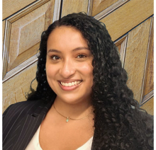 light skinned woman with long curly dark brown hair with black jacket and white shirt in front of wood paneling