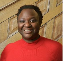 smiling dark skinned person with short curly black hair, red shirt in front of wood paneled wall