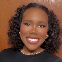 A head-and-shoulder picture of a person with shoulder length curly black hair. She is wearing a black top, looking at the camera, and smiling