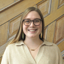 light skinned person with long blond hair, glasses, earrings, tan v-necked sweater in front of wood paneled wall,