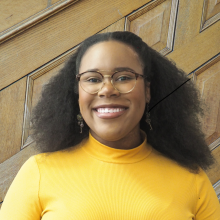 Head & shoulder picture of smiling person with brown skin, curly black hair, glasses and yellow shirt in front of wood paneled wall