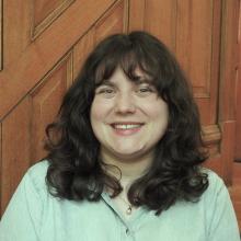 Head & shoulder picture of smiling person with light skin, way dark brown hair, with a light denim shirt in front of wood paneled wall