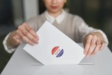 Image of a light skinned woman inserting a white envelope into a white voting box.