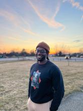 A dark skinned man with black glasses, a black crewneck sweatshirt, and a tan beanie.