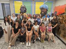 diverse group of people sitting on a couch with 2 rows of people standing behind them. behind this group is a blue and yellow mural featuring a dark-skinned man and woman