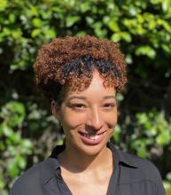 smiling person with curly brown hair gathered on top of head with a black shirt in front of blurry green trees