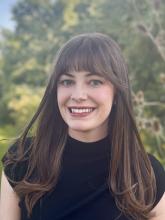 smiling light skinned person with long brown hair and bangs, green eyes in front of blurred greenery