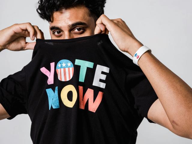 man pulling a black t-shirt that says "Vote Now" over his head