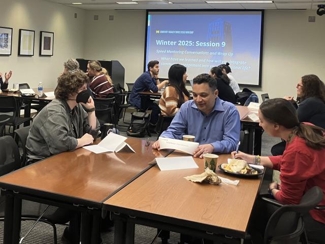 A group of twelve students sit at four tables conversing in groups of three.
