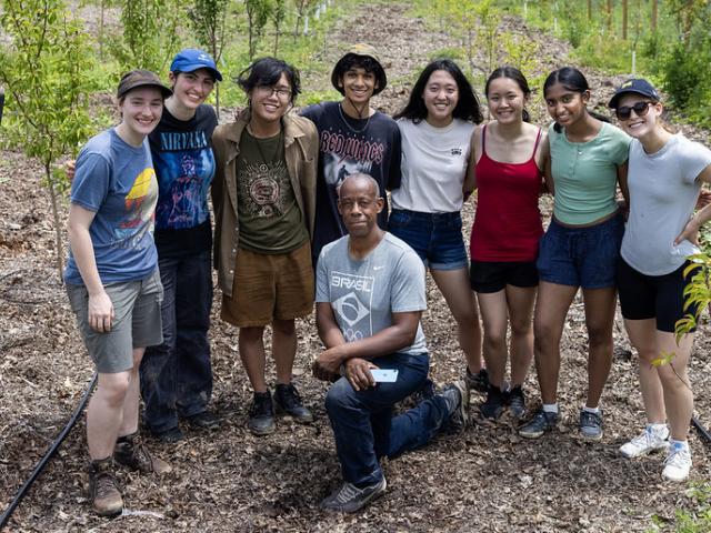 A group of students standing behind a man in the middle of a planted forest.