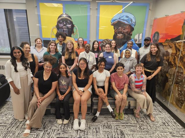 A group of students sit next to various pieces of artwork.
