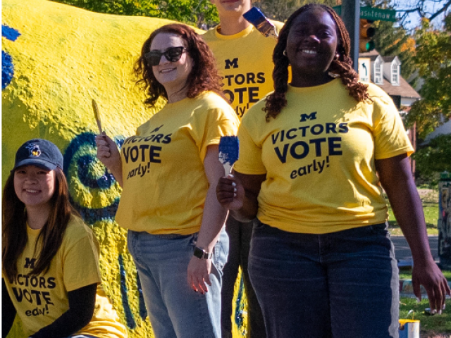 Four students wearing Victors Vote Early shirts standing in front of a yellow boulder.