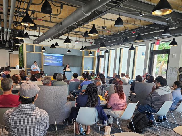 Rosalie Ginsberg Commons filled with people seated at chairs and tables facing 2 people speaking under large TV monitors. 
