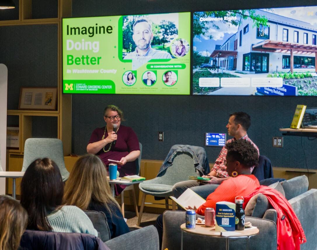 seated person holding a microphone under a digial screen with words 'Imagine Doing Better' looking at seated man in plaid shirt in front of an audience. 