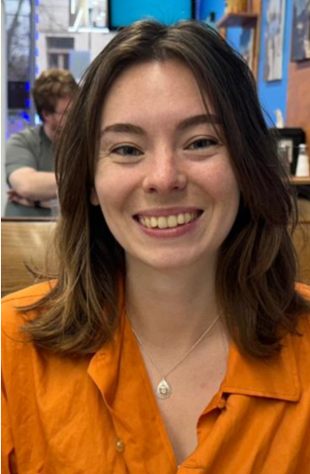 smiling light skinned person with shoulder-length brown hair with orange button-down shirt and pendant necklace